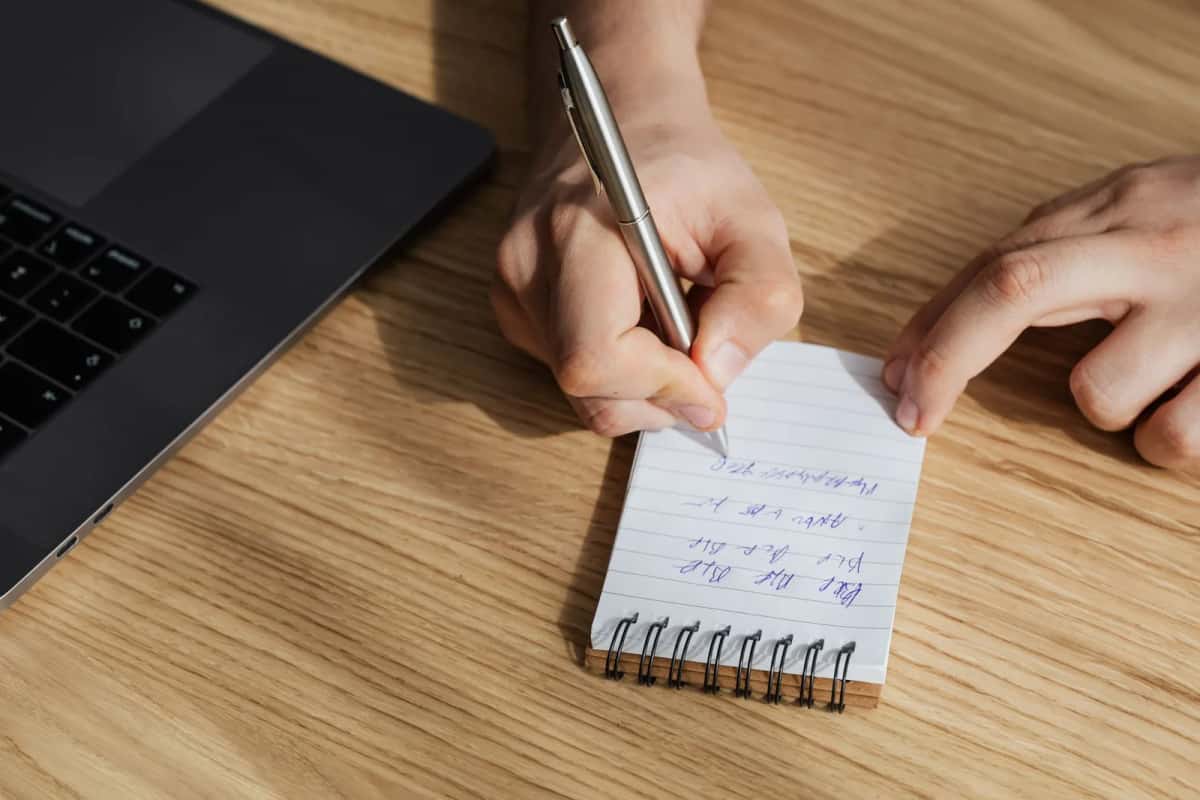 Une main prend des notes sur un carnet posé à côté d'un ordinateur portable