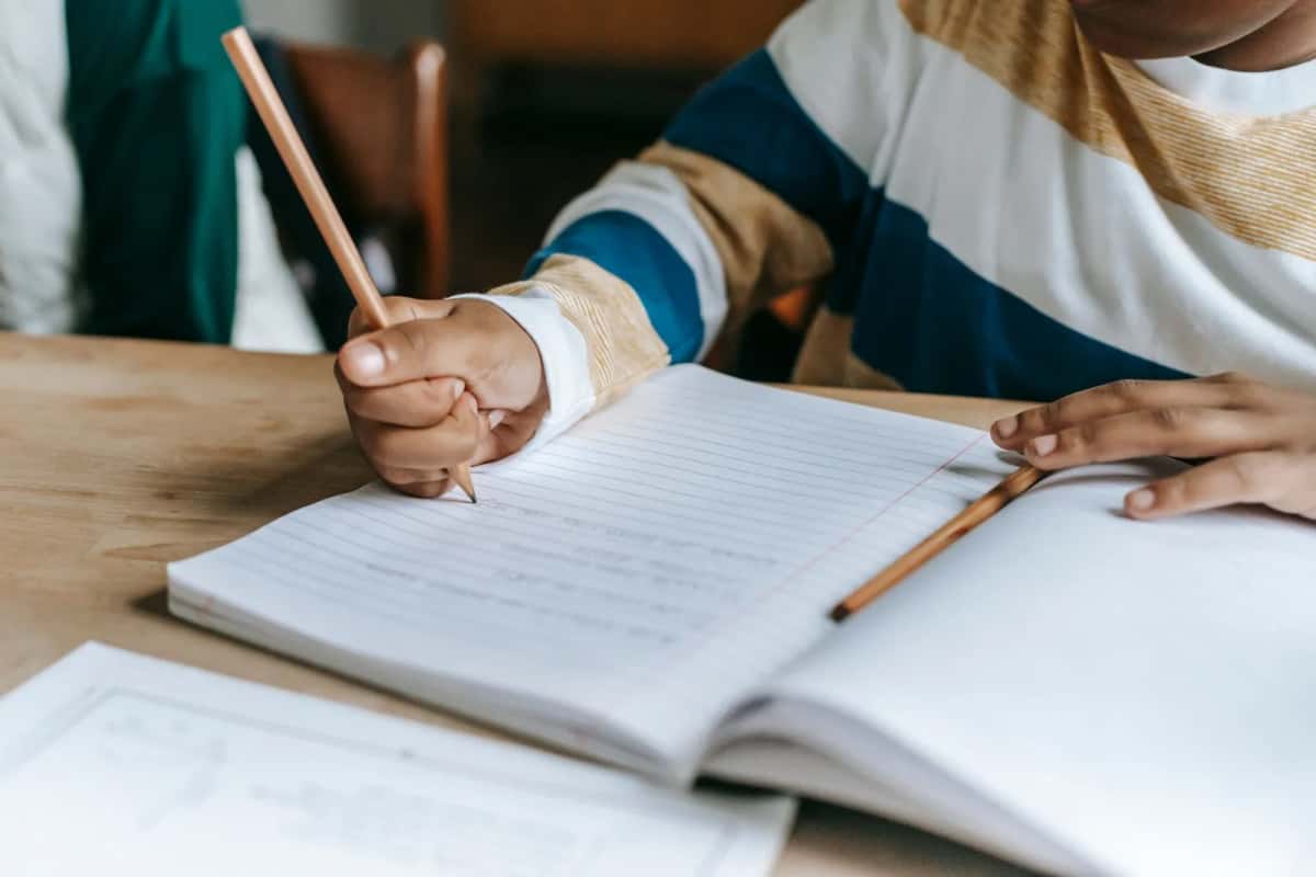 Enfant qui écrit au crayon dans un cahier posé sur une table en bois